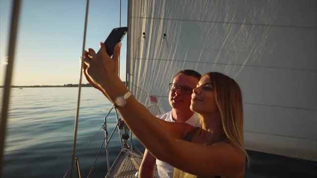 Girl and guy are taking self-portrait photo by phone on a yacht in evening. Woman is holding phone, they are smiling and posing on background of big sail.