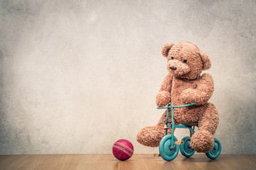 Teddy Bear riding on old retro toy tricycle and leather ball in front concrete textured wall...