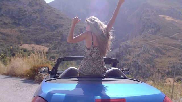 Slow motion close up shot of young blond female standing in convertible car on the edge of the cliff and raising her hands up against high rocky mountains.Happy girl enjoying success and stunning view