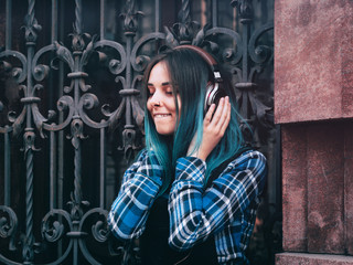 Street hipster girl listening music with brown headphones. Teen girl with blue dyed hair,piercing in nose,violet lenses,unusual hairstyle sitting near historic building.