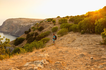 Among the fields and the sea, a tourist with a backpack enjoys the scenery at sunset and feels freedom, the view of the mountains can be seen from afar, the sun shines with bright rays.