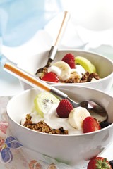 Muesli in two small porcelain bowls with yoghurt, fruit, caramel brittle, raspberries, strawberries, kiwi and a spoon