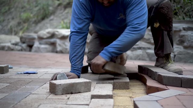 Very tight shot of stone mason fitting brick pavers into a herringbone patio design with an emblem in the middle.