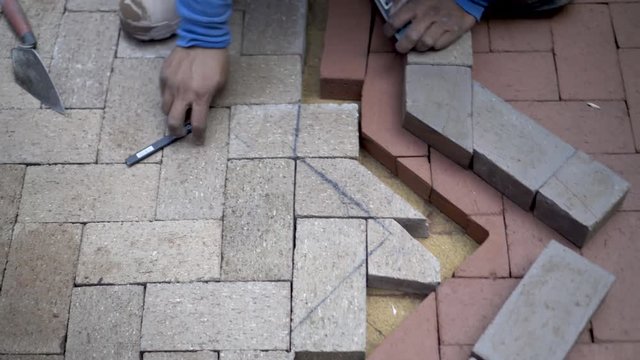 As camera turns and rotates around the action, a stone mason uses a level as a straight edge to mark bricks for cutting to fit a two-tone design.
