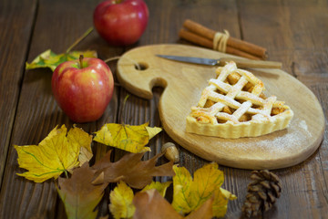 Piece of pie on a wooden board with a knife. Around with cinnamon sticks, apple, leaves, acorn, and...