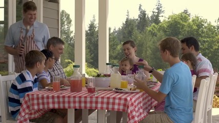 Man carries a T-bone steak in tongs to table of family and friends enjoying a BBQ together. Real time 4K.