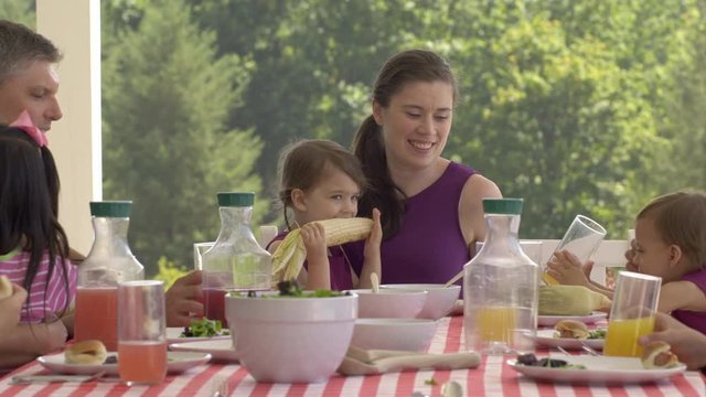 Young girl sits on mother's lap and eats sweet corn on the cob at table with family and friends enjoying a BBQ together. Real time 4K with camera dolly.