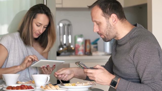 Couple with tablet computer and smartphone eating breakfast at home
