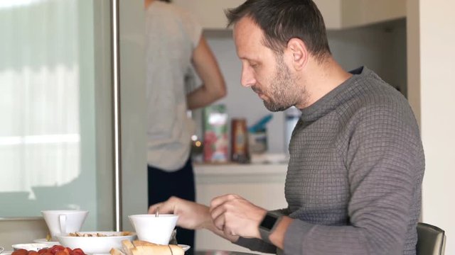 Man preparing tea during breakfast in kitchen at home, wife in the background
