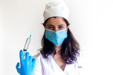 A female doctor with dark hair in a blue protective gloves and a mask holding a syringe filled with a vaccine. Vaccination against influenza. Injection, drip