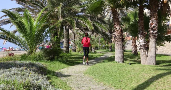 4k video. Back view of adult woman with red hoodie and black leggings, or knee pants, running on wooden footbridge, in park of beach in Benalmadena, Malaga, Andalusia, Spain
