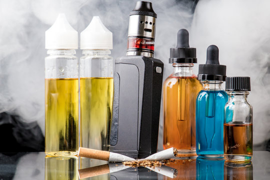 Tobacco cigarette crushed under electronic cigarette surrounded with e-juice bottles