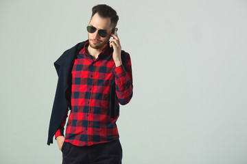 handsome young guy in a plaid shirt in sunglasses in the studio on a wall background talking on the...