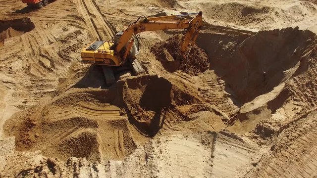 Excavator digging works in a quarry. Orange sand soil rock heavy industry heavy equipment. Aerial drone action flight around. Sunny day.