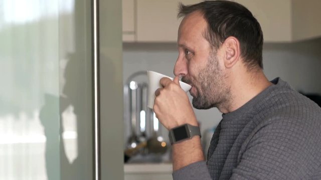 Young man watching TV and drinking coffee by table in kitchen at home
