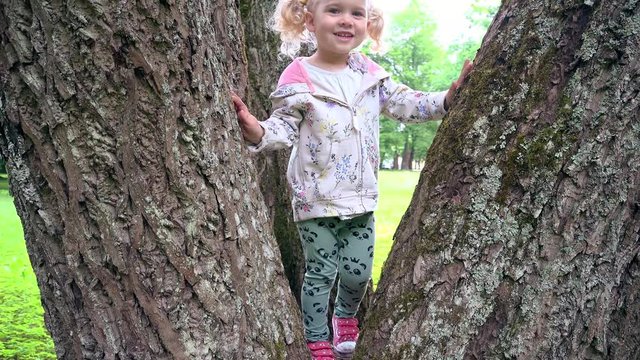 Cute blond girl playing between tree trunks in park