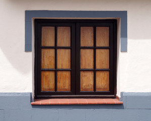 traditional old wooden window with glass panes closed internal shutters on a clean white house wall with gray painted surround and lower area in bright sunlight