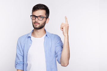 Waist up shot of joyful guy wearing blue long sleeve shirt and glasses looking up, pointing finger at copy space above his head. Bearded young man indicating something on blank studio wall with hand.