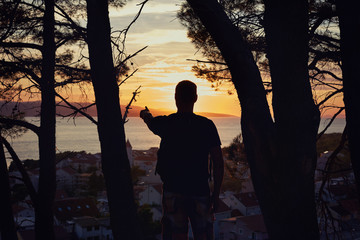 Fototapeta na wymiar Sunset side view of male tourist silhouette searching location pointing with the finger at panoramic view of beautiful Croatian landscape with rolling hills, gardens and houses of the ancient town.