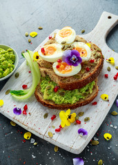Tasty sandwich with avocado boiled eggs, pumpkin seed and edible viola flowers in a white board. healthy food