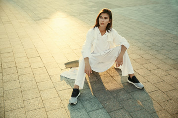 girl in a white suit is sitting on a longboard on the sunny square of the old city