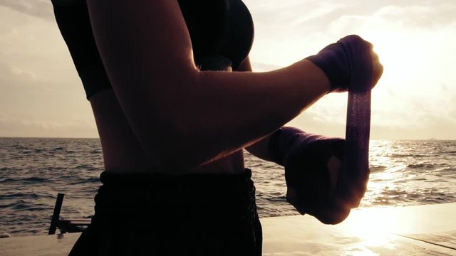 Young fit woman getting her fists ready for the boxing gloves by wrapping bandage around them standing against the sun. Female boxer wrapping hands with boxing wraps on the beach. Lens flare