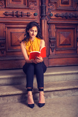 Way to Success. Power of Reading. East Indian American college student studying in New York, sitting on steps in front of vintage style library door way, looking down, reading red book, thinking..