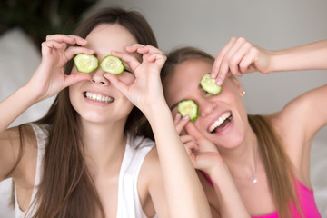 Two young smiling girlfriends being silly, putting cucumbers on their eyes and making funny faces. Best friends having fun, using natural product for beauty routine. Close up, facing camera portrait.