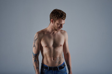Fashionable bearded European man with stylish tattoos on his fit muscular body posing isolated with naked torso, wearing blue jeans with black belt, looking down with serious facial expression