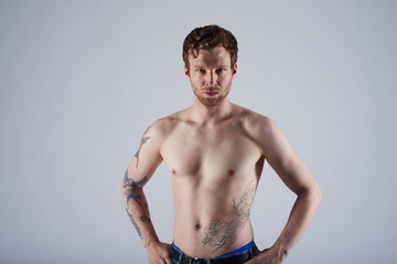 Studio portrait of handsome and confident Caucasian adult male model with stylish trimmed beard and tattooed body demonstrating his perfect sporty torso at camera, holding hands on his waist