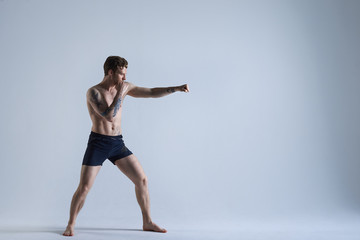 Full length shot of muscular athletic young Caucasian kickboxer training in studio, punching air, preparing for fight, wearing black shorts and no shoes. People, sports, boxing and fighting