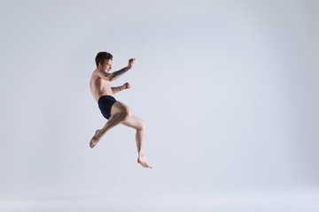 Fototapeta na wymiar Freeze action shot of flying shirtless and barefooted young male boxer wearing trunks jumping high against blank grey studio wall background with copy space for your advertising information