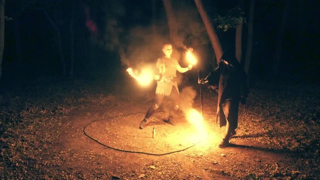 Fire show. Fiery performance. A girl in the mantle sets on fire a circle on the ground in the nocturnal forest, while another girl in chain mail performs a fiery performance inside the circle. 