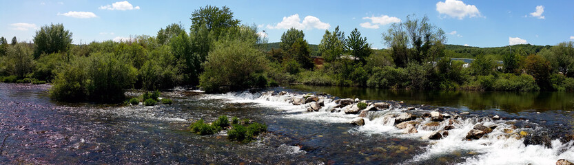 Naklejka na ściany i meble panoramic photo of a waterfall on a small river, with green trees on the opposite side