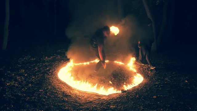 Gothic girl is performing a fiery show, standing in the middle of burning circle in the night forest. Fire performance