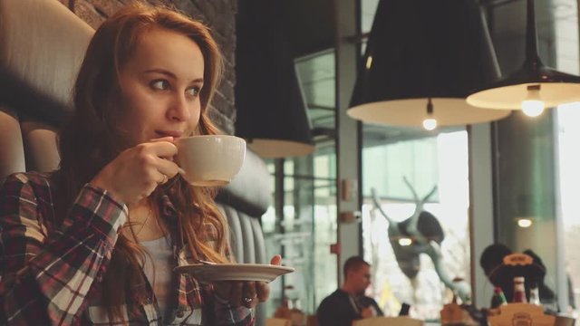 Woman drinking coffee in a cafe, 4K Slow Motion 120 fps. Stabilazed dolly shot. Young beautiful calm woman enjoying a cup of hot coffee in the restaurant. Relaxed morning breakfast in a city. 
