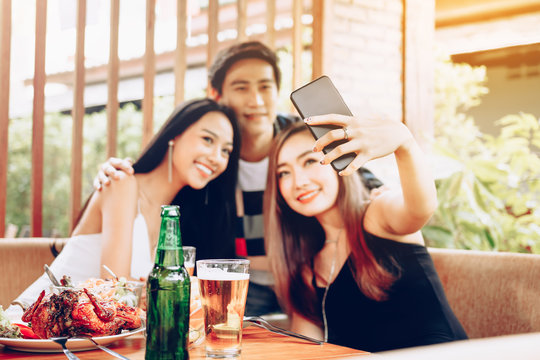 Group of asian friends sitting at bar restaurant and taking selfie with mobile smartphone.