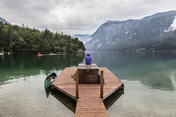 Solitary woman wearing purple hoodie watching tranquil overcast morning scene at lake Bohinj, Alps...