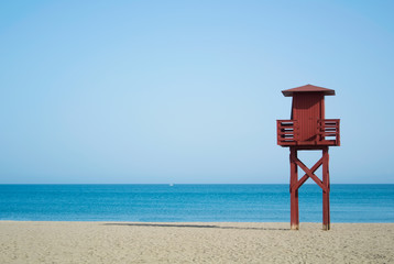 Red wooden lifeguard tower on the abandoned beach at Benalmadena, Malaga province, Spain. Beautiful view of the sea and sandy beach on sunny summer day with copy space. - obrazy, fototapety, plakaty