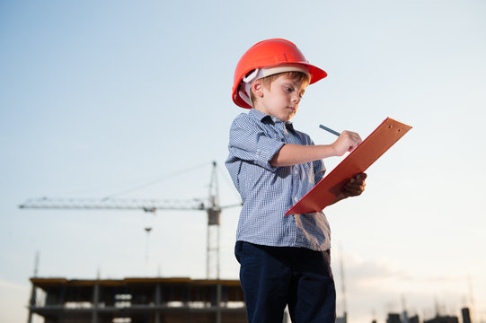 kid builder wearing orange helmet takes notes on building site background