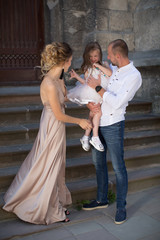 The young happy parents standing outdoor against the background of old building and father holding little cute daughter in his arms, young elegant mother looking at daughter