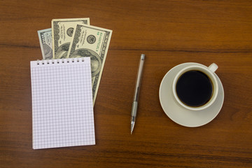Cup of coffee, dollars, notepad and pen on wooden desk