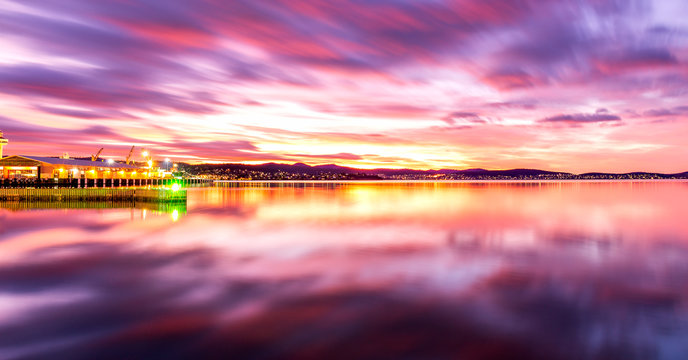 Pink sunrise on the Hobart pier (Tasmania) on a cloudy morning. Purple and fire in the sky reflecting on the very calm water on a windless day.