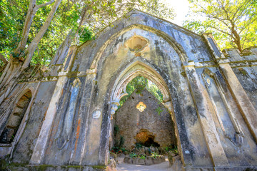 Ruins of chapel in the park of Monserrate, on the hills above Sintra, Portugal, Europe. Main facade of the ancient church covered with vegetation.