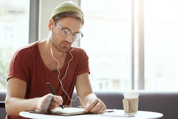 Picture of stylish young man freelancer of creative occupation writing down in copybook, noting...