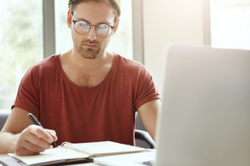 Horizontal image of handsome stylish young male entrepreneur making notes in copybook, sitting at cafe table in front of open laptop and waiting for partner to discuss business issues over coffee