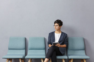 Horizontal portrait of young attractive woman being in anticipation of meeting with potential partners, sits on chair in waiting room, uses electronic tablet not to feel bored, looks pensively aside