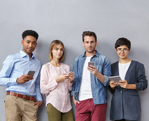 Friendly business partners wait for boss, discuss together project, share opinions with each other, use modern gadgets for illustaring ideas with schedules and diagrams, stand in one line at grey wall