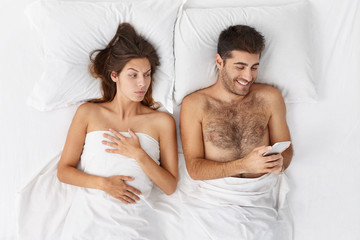 Obraz na płótnie Canvas Happy bearded male lying in bed reading message on phone from his lover and smiling broadly, his suspcious girlfriend lying next to him, trying to peek at screen. Cheating and infidelity concept