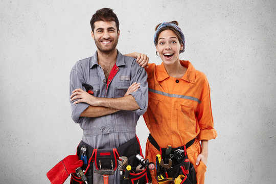 Happy excited female and male plumber wear uniform with tool belts, being glad to repair bathtub, recieve pleasant comments from clients, isolated over white concrete wall. Success, repairing concept